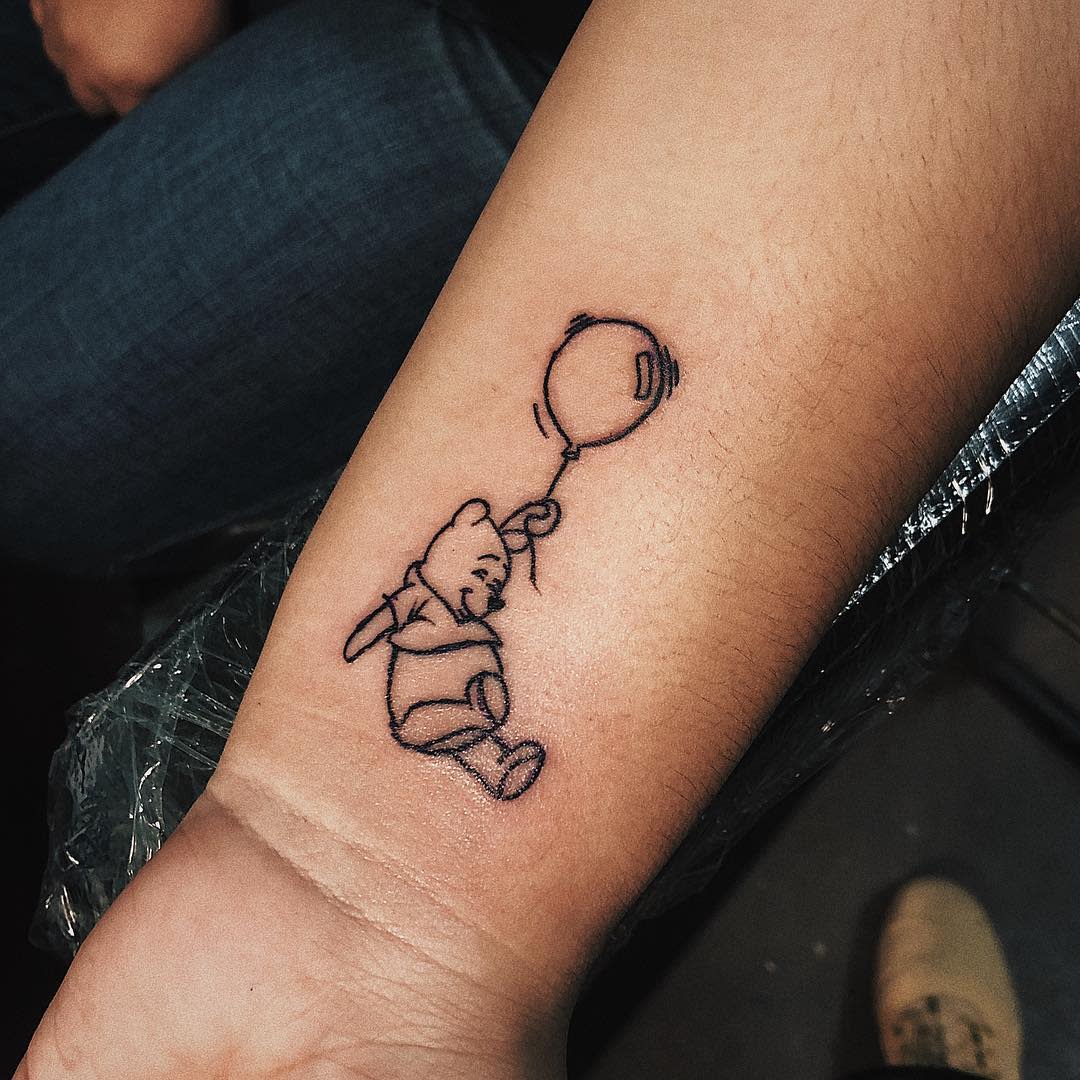 Simple outline tattoo of Winnie the Pooh holding onto a balloon on a person's wrist